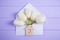 Beautiful white tulips in envelope with wooden icon of house with hole in form of heart on lilac wooden background