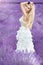 Beautiful woman with blond hair in a beautiful long white wedding dress stands on a field with heather flowers