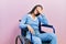 Beautiful woman with blue eyes sitting on wheelchair covering eyes with hand, looking serious and sad