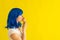 A beautiful woman with blue hair stands in profile and licks a horn of raspberry ice cream on a yellow background. The
