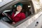 Beautiful woman in bright red coat, white woolen scarf and knit green hat smiles while driving a car. Female driver confidently