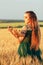 Beautiful woman in green dress on field with sheaf of wheat in hand at sunset light, girl enjoying summer nature landscape,