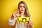 Beautiful woman holding amazed wow surprise banner over isolated yellow background pointing with finger to the camera and to you,