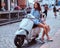 Beautiful woman with long brown hair dressed in trendy clothes posing with white retro Italian scooter on the street.
