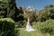 a beautiful woman with long brown hair and long white dress stay along a path along beautiful bushes in the park rear