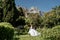 a beautiful woman with long brown hair and long white dress stay along a path along beautiful bushes in the park rear