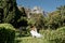 a beautiful woman with long brown hair and long white dress stay along a path along beautiful bushes in the park rear