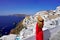Beautiful woman in Oia village holds hat when looking cityscape from terrace in Santorini Island, Greece