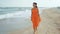 Beautiful woman in orange dress goes through the sea sandy beach and pensively looks forward