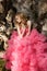 Beautiful woman in pink evening dress with fluffy aerial skirt is posing in botanical garden on the driftwood