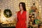 Beautiful woman in red dress with glass of champagne posing in decorated room with heap of christmas gifts