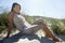 Beautiful Woman Sitting On Sand Dune At Grassy Beach