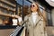 Beautiful woman in the sun of goggles in a beige raincoat and shopping bags in her hands, stands in front