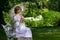 Beautiful woman in vintage dress sitting on ornate metal bench holding parasol next to river