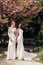 Beautiful women in luxurious wedding dresses with accessories posing in garden with blossoming sakura trees