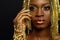 Beautiful young african woman posing at studio in golden jewellery, face with hand portrait over dark background