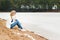 Beautiful young blonde girl in jeans and a white shirt sitting on the shore of the frozen cold of the lake near the forest