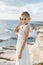 Beautiful young blonde model woman with nude makeup in a fashionable wedding dress walking at the sea coast at Cyprus