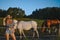 Beautiful young blonde woman dressed safari style in hat and plaid shirt posing with thoroughbreds horses on farm in