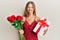 Beautiful young blonde woman holding anniversary present and bouquet of flowers sticking tongue out happy with funny expression