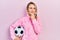 Beautiful young blonde woman holding soccer ball serious face thinking about question with hand on chin, thoughtful about