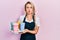 Beautiful young blonde woman wearing waitress apron holding take away cup of coffees puffing cheeks with funny face