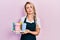 Beautiful young blonde woman wearing waitress apron holding take away cup of coffees relaxed with serious expression on face