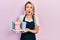 Beautiful young blonde woman wearing waitress apron holding take away cup of coffees in shock face, looking skeptical and