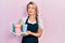 Beautiful young blonde woman wearing waitress apron holding take away cup of coffees smiling looking to the side and staring away