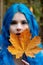 Beautiful young bohemian woman with blue hair happily holding an autumn leaf in her hands