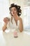 Beautiful young brunette woman in hair curlers taking cream from cupcake with her finger sitting with cup of coffee in bright