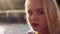 Beautiful young caucasian girl with blond hair and red lips smiles sitting in front of water on sunset. Looking in to