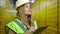 Beautiful young Caucasian woman in hard hat thinking filling in paperwork standing in yellow warehouse. Portrait of