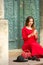 Beautiful young caucasian woman sitting on the stairs by the door with travel suitcase and smartphone wearing long red