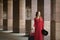 Beautiful young caucasian woman wearing red long dress with a suitcase standing in columns.