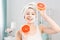 Beautiful young Caucasian woman with white cleansing mask on half of face and grapefruit halfs on light background