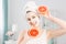 Beautiful young Caucasian woman with white cleansing mask on half of face and grapefruit halfs on light background