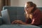 A beautiful young charming girl with glasses lies on her stomach  on a blue sofa in her living room and works on a laptop, distant