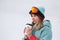 A beautiful young chewing woman in an extreme sports suit, hat and helmet with a mask on the winter slope is holding a