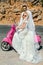 Beautiful young couple, blonde female model with her husband in amazing wedding clothes sitting on scooter