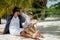 A beautiful young couple joyfully sits on white sand under a palm tree on the seashore under a green palm tree. Honeymoon Travel