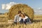 Beautiful young couple in love in casual clothes sitting in field near hay bale. Ecotourism in countryside concept.