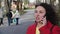 Beautiful young curly girl in a red coat is chatting on a smartphone in the city