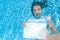 Beautiful young girl holding white blank board in swimming pool under water, fun on family vacation