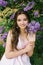 Beautiful young girl sitting on the ground in the garden with blooming lilac. She is happy and smiles a beautiful smile with white