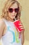 Beautiful young girl in sunglasses in the summer warm day drinking coke through a straw with red glass