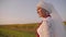 A beautiful young girl in a white national dress stands in the field and looks towards the sunset and then turns around to the cam