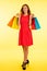 Beautiful young haapy woman in vibrant red dress holding shopping bags over yellow background