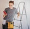 Beautiful young happy woman repairman standing by a stepladder in a belt with tools on a white wall background