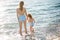 Beautiful young mother and daughter having fun resting on the sea. They stand in the water in the same swimsuit, back in the frame